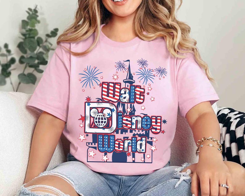 May include: A light pink t-shirt featuring the text "Walt Disney World" in red, white, and blue, with a castle and fireworks graphic. The shirt has short sleeves and is made of a soft material.