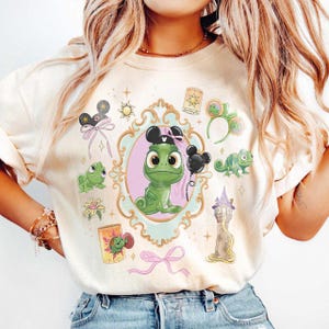 Könnte beinhalten: Ein cremefarbenes T-Shirt mit einer zentralen Grafik eines grünen Chamäleons mit schwarzen Mausohren in einem verzierten goldenen Rahmen. Um den Rahmen herum befinden sich Illustrationen eines Turms, einer Laterne, einer Blume und grüner Chamäleons.