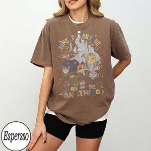 May include: Brown t-shirt with a colorful graphic featuring cartoon characters and the text "WHERE ANYONE CAN BE ANYTHING." The shirt has short sleeves and a relaxed fit. The model is wearing black shorts and a white undershirt.