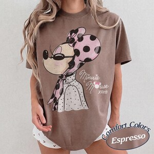 May include: Espresso-colored t-shirt featuring a vintage-style Minnie Mouse graphic. Minnie is depicted with a pink polka-dot bow and scarf, wearing sunglasses. The shirt has the words "Minnie Mouse XOXO" printed on it. The shirt is a Comfort Colors brand.