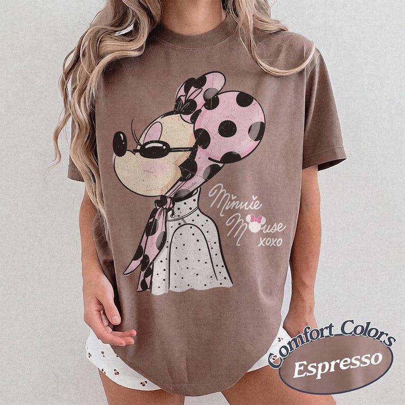 May include: Espresso-colored t-shirt featuring a vintage-style Minnie Mouse graphic. Minnie is depicted with a pink polka-dot bow and scarf, wearing sunglasses. The shirt has the words "Minnie Mouse XOXO" printed on it. The shirt is a Comfort Colors brand.