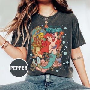 Pu&ograve; includere: T-shirt grigio scuro con una grafica colorata di Ariel de La Sirenetta. Il design include Ariel, Flounder ed elementi floreali. Il testo "THE LITTLE MERMAID" &egrave; visualizzato sopra i personaggi. La maglietta ha un aspetto vintage e usurato.