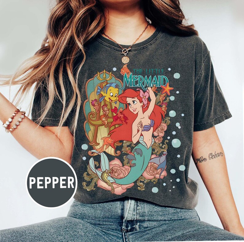 Pu&ograve; includere: T-shirt grigio scuro con una grafica colorata di Ariel de La Sirenetta. Il design include Ariel, Flounder ed elementi floreali. Il testo "THE LITTLE MERMAID" &egrave; visualizzato sopra i personaggi. La maglietta ha un aspetto vintage e usurato.