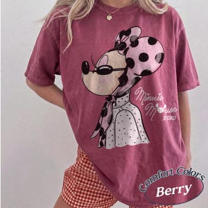 May include: Berry-colored t-shirt featuring a Minnie Mouse graphic. The design includes Minnie's face with sunglasses, a polka-dot bow, and the words "Minnie Mouse xoxo." The shirt is a Comfort Colors brand.