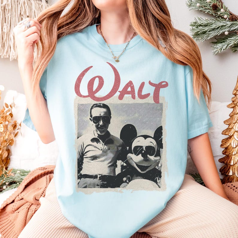 May include: Light blue t-shirt featuring a vintage-style graphic of Walt Disney with Mickey Mouse wearing sunglasses. The word "Walt" is printed above the image in a distressed red font. The shirt is a casual, comfortable style.