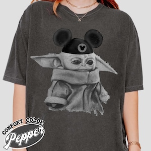 May include: A charcoal grey short-sleeve t-shirt featuring a graphic of a small creature with large ears wearing a black hat with mouse ears. The graphic is rendered in a sketch-like style. The text COMFORT COLOR Pepper is printed in the bottom corner.