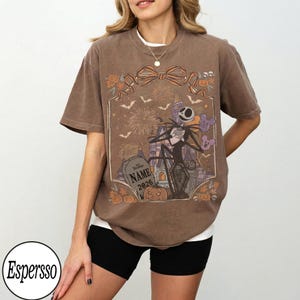 K&ouml;nnte beinhalten: Braunes T-Shirt mit einem Aufdruck von Jack Skellington aus The Nightmare Before Christmas. Das Design umfasst einen Grabstein, Flederm&auml;use, K&uuml;rbisse und den Text "NAME 2026". Das Shirt hat einen Vintage-Look.