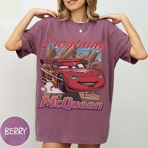 K&ouml;nnte beinhalten: Beerenfarbenes T-Shirt mit einer Grafik von Lightning McQueen aus dem Film Cars. Das Design enth&auml;lt den Text "Lightning McQueen" in Rot und "McQueen" in Lila. Das Shirt hat einen Vintage-Look.
