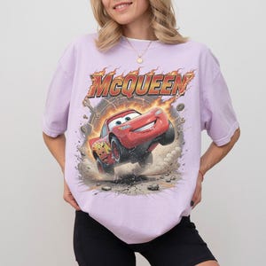 Op de afbeelding: Een lichtpaars t-shirt met korte mouwen en een grafische print van de rode raceauto Lightning McQueen. Het ontwerp bevat de tekst MCQUEEN in dikke gele en oranje letters, omringd door vlammen, mechanische tandwielen en stofwolken.