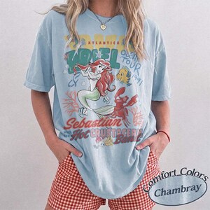 May include: Light blue t-shirt featuring a graphic of Ariel from The Little Mermaid, with the text "Ariel Ocean Tour 1991" and "Sebastian with the Hot Crustacean Band." The shirt is a Comfort Colors Chambray.