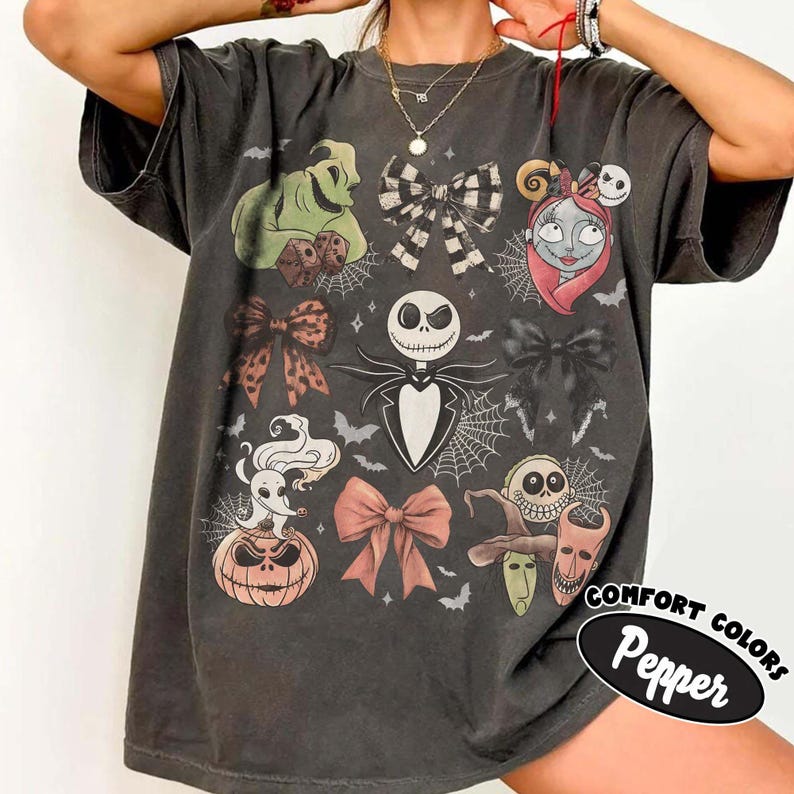 May include: A charcoal gray short-sleeve t-shirt featuring a graphic print of characters and bows from The Nightmare Before Christmas, including Jack Skellington, Sally, Zero, and Oogie Boogie, with the text Comfort Colors Pepper.
