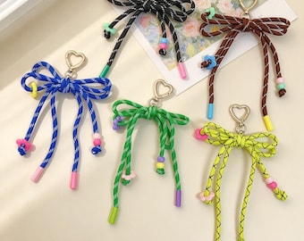 May include: Five decorative bow keychains made from patterned rope in black, brown, blue, green, and neon yellow. Each bow features a gold-tone heart-shaped clasp, small colorful flower-shaped beads, and solid-colored plastic end caps.