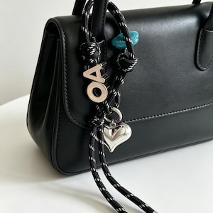 Può includere: Una borsa nera con un accessorio decorativo attaccato al manico. L'accessorio è composto da una corda nera e bianca maculata con un ciondolo a forma di cuore argentato, una lettera O argentata, una lettera A argentata e una piccola perla blu.