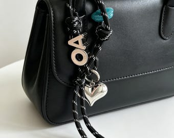 May include: A black handbag featuring a decorative accessory attached to the handle. The accessory consists of a black and white speckled rope with a silver-toned heart charm, a silver-toned letter O, a silver-toned letter A, and a small blue bead.