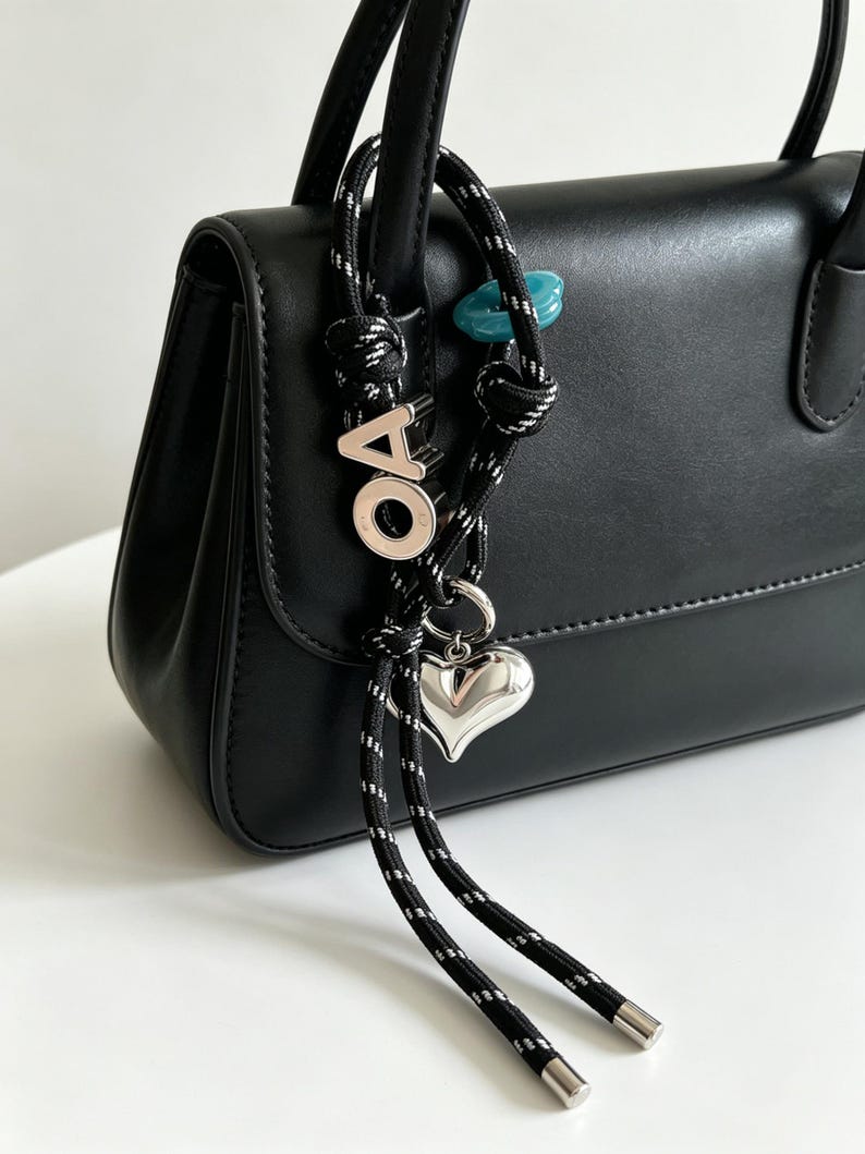 May include: A black handbag featuring a decorative accessory attached to the handle. The accessory consists of a black and white speckled rope with a silver-toned heart charm, a silver-toned letter O, a silver-toned letter A, and a small blue bead.