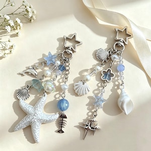 Puede incluir: Dos llaveros plateados en forma de estrella con dijes colgantes que incluyen estrellas de mar blancas, conchas marinas, un delfín, una espina de pescado y una estrella de cuatro puntas. Los diseños incorporan cuentas azules, transparentes y perlas.