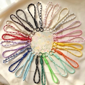 May include: A collection of twenty-one wrist lanyards arranged in a circular pattern. Each lanyard features a looped cord with a knotted design and a small metal ring at the base. The cords come in various solid colors and patterns, including black, white, red, blue, green, yellow, orange, purple, and pink.
