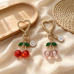 Puede incluir: Dos llaveros de metal dorado con cierres en forma de corazón y dijes de cerezas con hojas de esmalte verde. Un llavero tiene cuentas de cereza rojas translúcidas y un dije circular blanco con una O, el otro tiene cuentas rosas y una X.