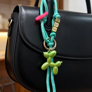 May include: A turquoise cord bag charm featuring a glossy lime green balloon dog pendant, a bright pink ring, and three gold-toned cube beads with the letters C, B, and X printed on them, all attached to a gold-toned metal ring.