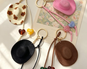 May include: Four miniature cowboy hat keychains with gold-tone metal rings and chains. One hat is cream with brown flowers, one is bright pink, one is black, and one is brown. Each keychain features two dangling heart-shaped beads in various colors.