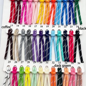 May include: A color chart featuring thirty-six numbered pairs of nylon cords, each with a reflective white diagonal stripe. The cords are arranged in three rows on a white display card, showcasing a variety of colors including red, pink, yellow, blue, green, purple, and black.