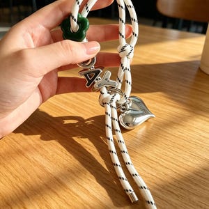 May include: A white rope keychain with black flecks featuring a dark green flower-shaped bead, silver-toned letter beads spelling out DAY, and a polished silver-toned heart charm. The rope ends are finished with smooth silver-toned metal caps.