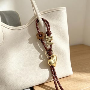 May include: A cream-colored tote bag with a textured surface features a brown rope charm attached to its handle. The charm includes a brown flower bead, a gold-toned letter M, and a polished gold-toned heart-shaped pendant hanging from a ring.
