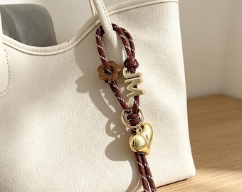 May include: A cream-colored tote bag with a textured surface features a brown rope charm attached to its handle. The charm includes a brown flower bead, a gold-toned letter M, and a polished gold-toned heart-shaped pendant hanging from a ring.