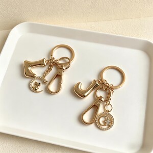 Puede incluir: Dos llaveros dorados, uno con la letra "J" y otro con el número "7". Cada llavero tiene un mosquetón y dijes decorativos, incluyendo una luna creciente con un conejo y una estrella dentro de un círculo, ambos adornados con pedrería.