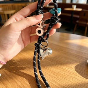 May include: A black rope lanyard with white reflective stitching featuring a silver-toned heart charm, a teal flower-shaped bead, and a silver-toned letter charm with the letters OA. The rope ends are finished with silver-toned metal caps.