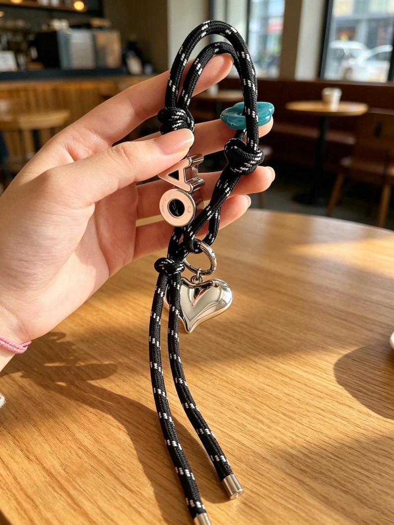 May include: A black rope lanyard with white reflective stitching featuring a silver-toned heart charm, a teal flower-shaped bead, and a silver-toned letter charm with the letters OA. The rope ends are finished with silver-toned metal caps.