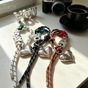 May include: Three rope keychains in white, black, and brown, each featuring a silver-toned metallic heart charm, a silver letter block, and a small colored plastic ring. The white keychain displays the text DAY, while the others show partial letters.