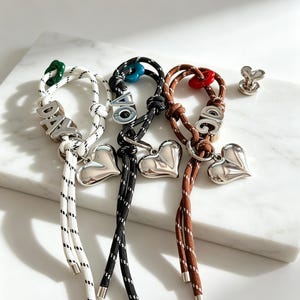 May include: Three cord bracelets in white, black, and brown, each featuring a silver-toned heart charm, a metal letter bead, and a small colored accent bead. The white cord has the letters DAY, the black has OA, and the brown has OG.
