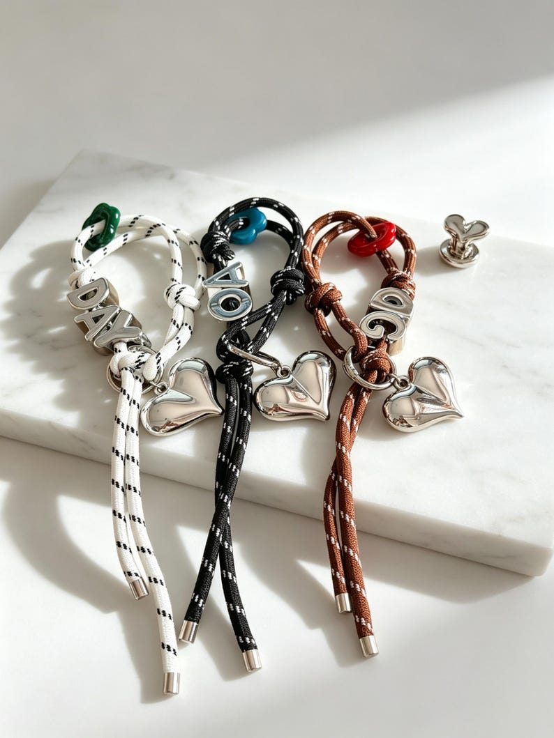May include: Three cord bracelets in white, black, and brown, each featuring a silver-toned heart charm, a metal letter bead, and a small colored accent bead. The white cord has the letters DAY, the black has OA, and the brown has OG.