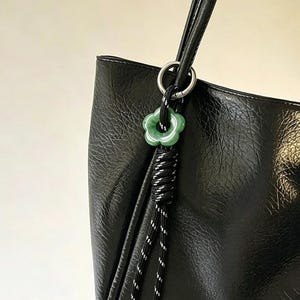 May include: A black textured tote bag featuring a decorative charm attached to a silver ring. The charm consists of a green flower-shaped bead threaded onto a black and white speckled cord that hangs down in two separate strands with metal tips.