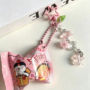 Pode incluir: Um chaveiro rosa com um pacote em miniatura de macarrão instantâneo. O chaveiro tem um fecho em forma de estrela, uma corrente de contas e pingentes decorativos, incluindo um peixe, flores e estrelas. O pacote de macarrão tem uma ilustração de um frango de desenho animado.