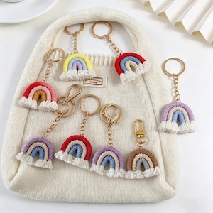 May include: A collection of rainbow-shaped keychains in various colors, including red, yellow, purple, and brown. Each keychain features a gold-colored chain and a white fringe at the bottom. The keychains are displayed on a white bag.