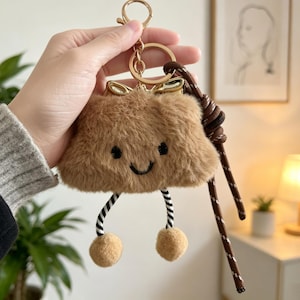 May include: A brown plush keychain with a smiling face, black and white striped legs, and two tan pom-poms. The keychain has a gold-colored ring and a brown rope detail.