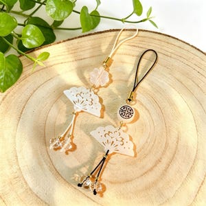 May include: Two white fan-shaped charms with intricate cut-out designs, hanging from gold-colored hardware. One charm has a pink flower accent, the other a white bead with a black symbol. Clear crystal beads dangle from the fans.