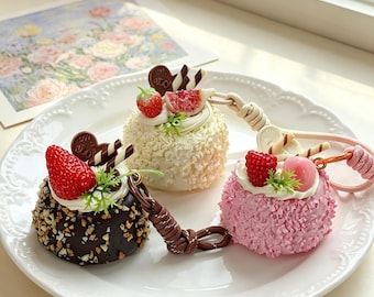 May include: Three miniature cake-shaped keychains on a white plate. One is dark brown with nuts, one is white with shaved chocolate, and one is pink. Each features a dollop of white cream, a piece of fruit, a chocolate heart, and wafer sticks.