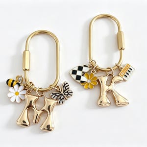 May include: Two gold-toned oval carabiner keychains. The first features a letter M charm, a white daisy, a bee, and a butterfly. The second features a letter K charm, a black and white checkered heart, a yellow daisy, and a piano keyboard charm.