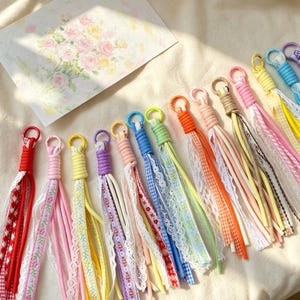 May include: A collection of multi-coloured fabric tassels featuring circular metal rings at the top. Each tassel is constructed from various ribbons, lace, and gingham fabric strips in shades of red, pink, yellow, purple, blue, green, and orange.