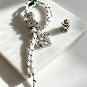 May include: A white rope bracelet with black flecks featuring silver-toned metal beads spelling the word DAY, a polished silver heart charm, and a small green accent bead. A separate small silver-toned metal object sits nearby.
