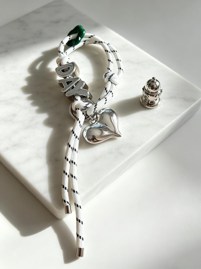 May include: A white rope bracelet with black flecks featuring silver-toned metal beads spelling the word DAY, a polished silver heart charm, and a small green accent bead. A separate small silver-toned metal object sits nearby.
