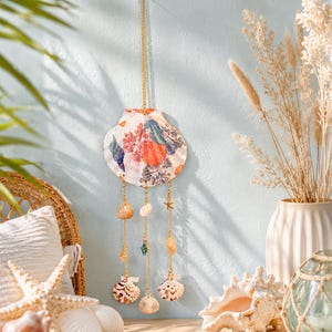May include: A decorative wall hanging featuring a large central scallop shell with a colorful floral and fruit print. Three gold-toned chains hang from the shell, each adorned with smaller natural seashells, a tiny gold starfish, and a blue bead.