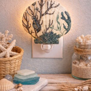 May include: A seashell-shaped night light with a detailed underwater scene in blue and green. The light is plugged into a white outlet. The image also includes a basket of starfish, sea glass, and a jar filled with sand and shells.