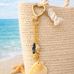 May include: A gold-toned heart-shaped lobster clasp keychain featuring three delicate gold chains adorned with a small dark blue seahorse charm, a tiny spiral seashell, and a larger, fan-shaped cream and yellow scallop shell pendant.