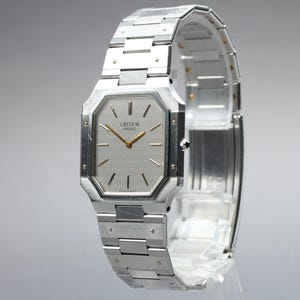 May include: A silver-toned Credor Seiko watch with a rectangular face and a matching metal band. The watch face is light gray with gold-colored hands and markers. The band is composed of rectangular links.