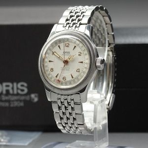 Puede incluir: Un reloj de pulsera Oris Big Crown Pointer Date plateado con esfera blanca texturizada, números arábigos y brazalete de eslabones de acero inoxidable. El reloj cuenta con una aguja central de fecha con una punta de media luna roja.