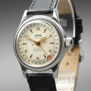 Puede incluir: Un reloj de pulsera automático Oris con bisel estriado plateado, esfera color crema con centro texturizado, manecillas doradas y números arábigos. El reloj incluye una manecilla de fecha, una correa de cuero negro y el texto Swiss Made.